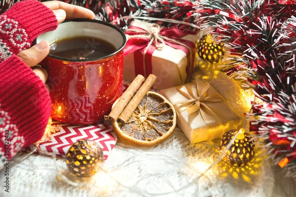 Fototapeta A woman's hand in a warm sweater holds a red mug with a hot drink on a table with Christmas decorations. New year's atmosphere, cinnamon sticks and a slice of dried orange, gifts, garland and tinsel