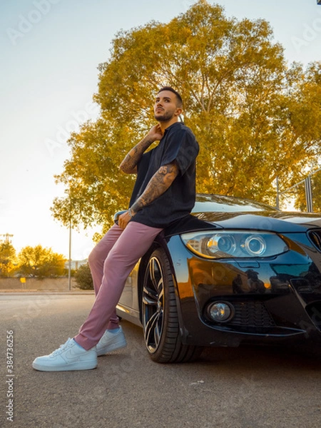 Obraz Hombre joven con tatuajes apoyado en su coche durante la puesta de sol