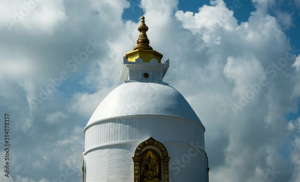 Fototapeta The upper part of the rounded Buddhist Peace Stupa, with a golden spire, against a background of white clouds