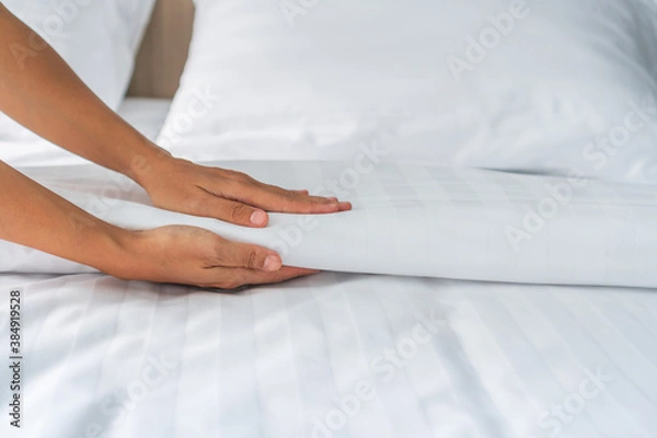Fototapeta Close up of maid hands set up white bed sheet in hotel room, copy space for text
