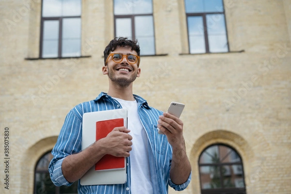 Fototapeta Portrait of young handsome Indian student holding laptop, book, mobile phone, walking to university. Smiling asian business man using smartphone, working online. Successful business