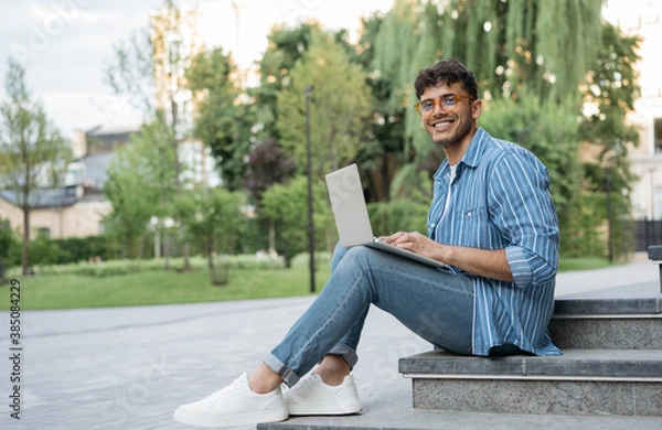 Fototapeta Portrait of happy Indian programmer using laptop computer, internet, working freelance project online, sitting in park. Asian student studying, learning language, using modern technologies outdoors