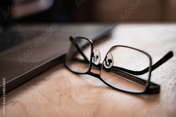 Fototapeta Background image of business office with laptop on wooden desk, focus on black hard rim glasses in foreground, copy space. Out of focus. Blurred