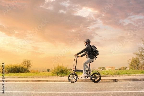 Fototapeta Smiling man with helmet and his electric bicycle at sunset. Commuter and new transportation concept. Green energy. Caucasian man drives his foldable e bike to go work or in leisure
