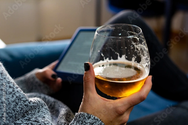 Fototapeta A woman with painted nails, holding a glass of beer in the foreground, can be seen in the background holding an e-book with the other hand. All of this while she rests on an armchair at home