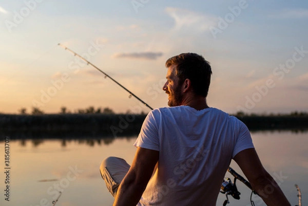 Fototapeta Back view of fisherman sitting at the river dock and fishing at sunset.