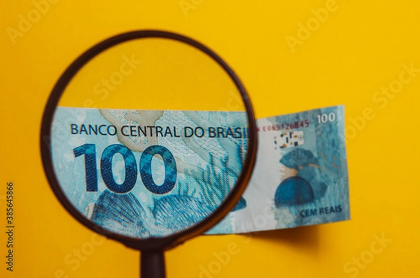 Obraz Brazilian money being viewed through a magnifying glass (in Brazil: LUPA), with notepad and calculator. Finance and salary concept. Economy of Brazil. Yellow background