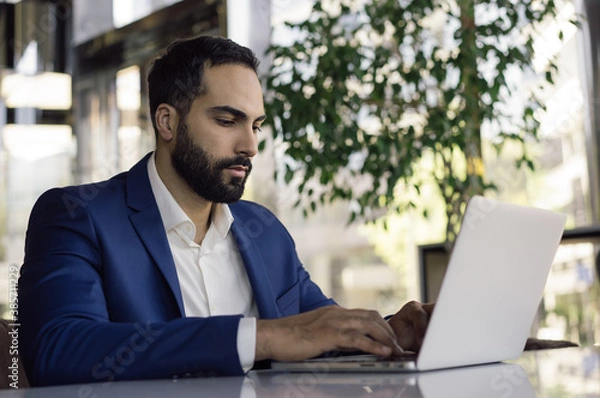 Fototapeta Young handsome arabic businessman planning project, typing on keyboard in modern office. Portrait of pensive middle eastern man wearing formal clothes, working online, sitting at workplace