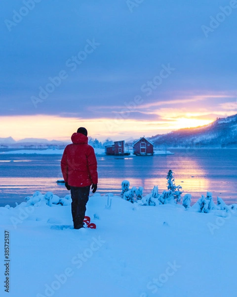 Fototapeta A man with snowshoes under moody sky winter sunset blue hour over the red houses in Norway