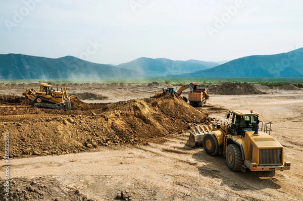 Fototapeta Earthworks in an open area on a summer sunny day. Cargo dump trucks, excavators, wheel loaders and bulldozers operate at the same time.