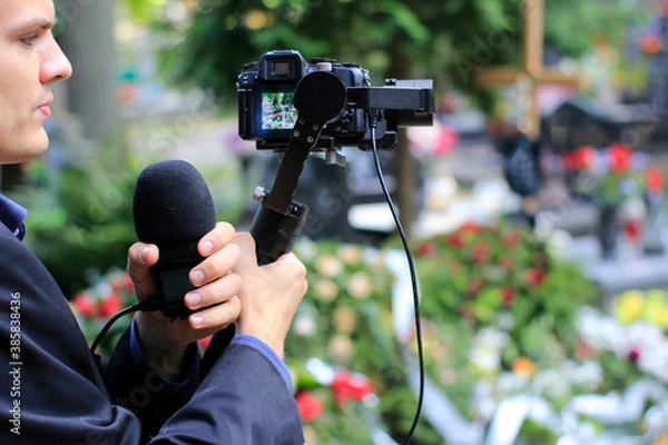 Obraz Elegant man in suit is recording grave with cross full of flowers. Funeral live streaming on cementary. Cameraman handing a microphone and camera on a gimbal.