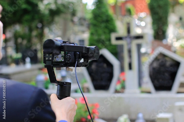 Obraz Elegant man in suit is recording grave with cross full of flowers. Funeral live streaming on cementary. Cameraman handing a microphone and camera on a gimbal.