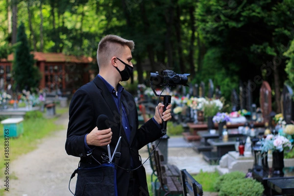 Obraz Elegant man in suit is recording grave with cross full of flowers. Funeral live streaming on cementary. Cameraman handing a microphone and camera on a gimbal.