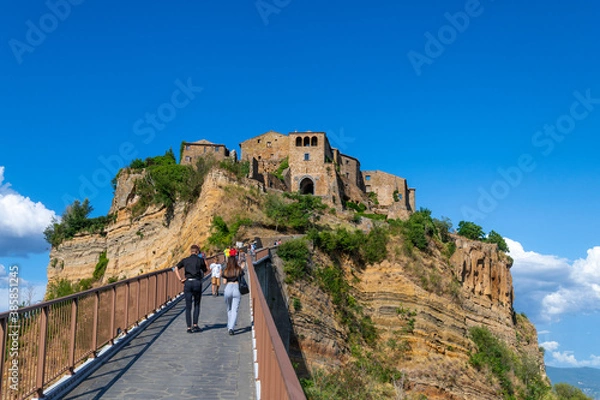 Fototapeta Civita di Bagnoregio. A young couple is crossing the bridge to reach the town set in the clouds, known as The Dying Town, on the top a plateau of volcanic tuff, Lazio Italy.