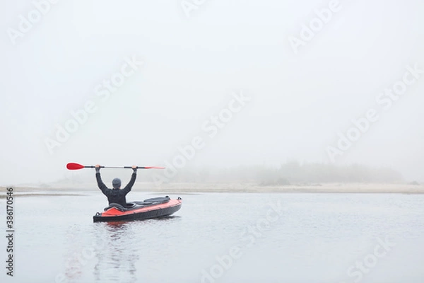 Fototapeta Male padding in river padding, sitting in canoe with raising paddle, enjoying water sport and beautiful nature, wearing black jacket and gray cap.