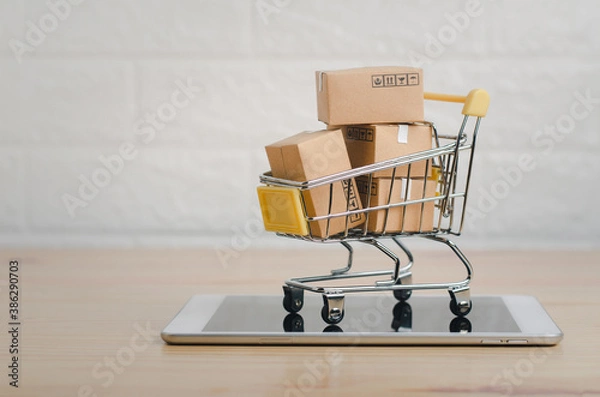 Fototapeta Brown paper boxs in  a shopping cart with tablet on wood table in office background.Easy shopping with finger tips for consumers.Online shopping and delivery service concept.