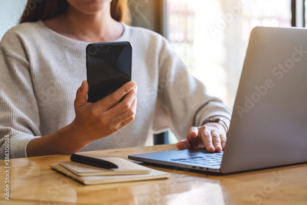Fototapeta Closeup image of a woman holding and using mobile phone and laptop computer in office