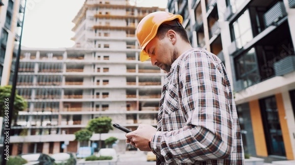 Fototapeta Young Engineer using smartphone on construction site. Architect using phone. Manager development building texting on smart phone. Builder makes notes in application during inspection and verification