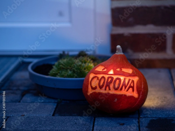 Fototapeta A carved illuminated pumpkin with corona lettering laying in front of a house entrance