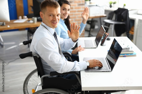 Obraz Businessman in wheelchair and businesswoman at table are smiling and raised their hands in greeting. Positive work mood in people with disabilities concept