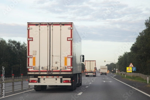 Fototapeta European heavy trucks with white semi trailers van drive on left side of two lane suburban asphalted highway road, back view at summer evening on forest and sky background, transportation logistics