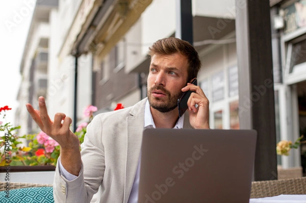 Fototapeta The upset businessman is working on the laptop and talking on his phone in the coffee bar 