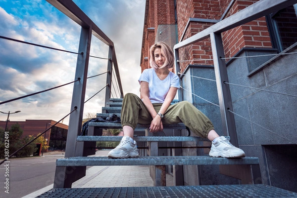 Fototapeta Girl in hip hop clothes sitting on a metal staircase looking at camera with a rebel look in an urban environment at sunset
