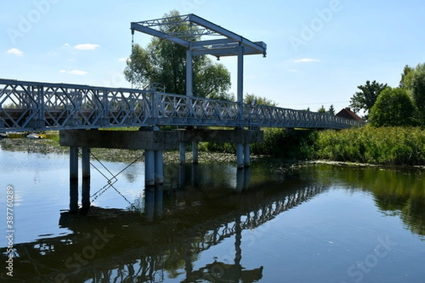 Fototapeta Close up on a metal pedestrian and car bridge connecting two river banks or coasts with one of the coasts being overgrown with shrubs, reeds, and other types of flora seen on a sunny summer day