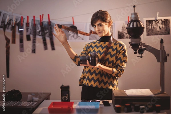 Obraz Happy young female holding filmstrip and smiling. Portrait of creative girl photographer in photo studio darkroom. Developing analog camera film