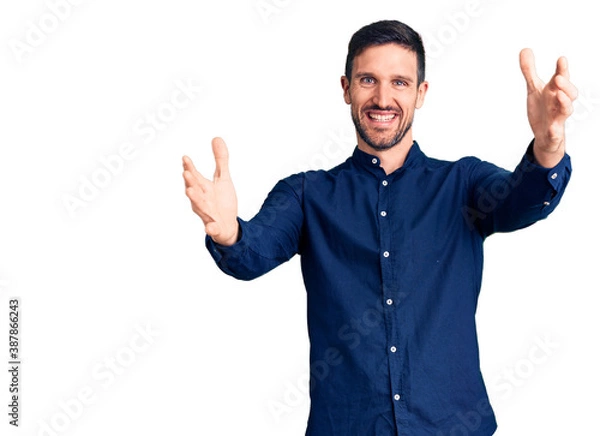 Fototapeta Young handsome man wearing casual shirt looking at the camera smiling with open arms for hug. cheerful expression embracing happiness.