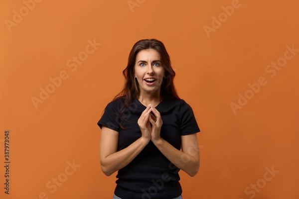 Fototapeta It is for me Joyful caucasian woman in black t-shirt keeps hands at the chest, in anticipation of a pleasant surprise, happy to receive awesome present. Isolated over an orange background.