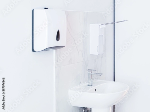 Fototapeta Bathroom with soap dispenser, white sink with metal faucet, and plastic tissue paper box, towel hanging on white wall. Light interior