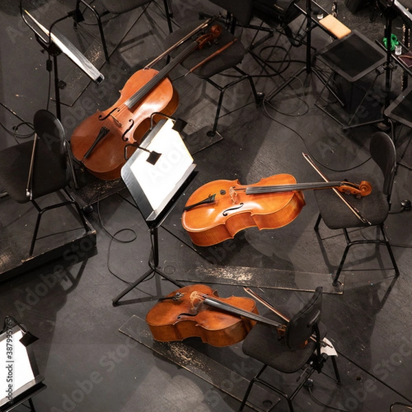 Fototapeta three cellos in the orchestra pit among the chairs and sheet music