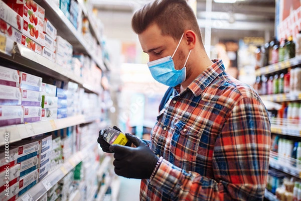 Fototapeta Young man is choosing a toothpaste at a supermarket wearing a medical sterile mask during the coronavirus pandemic quarantine. Healthcare, hygiene and home sanitizing concept.