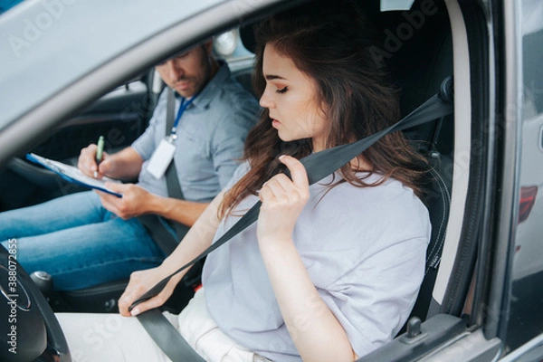 Fototapeta Beautiful young woman upset with results of driving tests. Strict examiner holds clipboard and makes notes of her mistakes. Female student and male instructor sit in a car with open window.