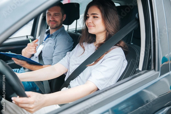 Fototapeta Male instructor satisfied with the ride of his female student, he smiles and comments on her correct actions. Two people are riding in a gray car and wearing safety belts.