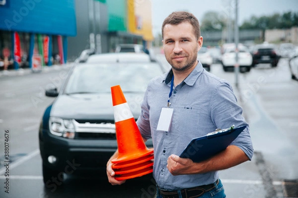 Fototapeta Male driving instructor standing with orange traffic cones and clipboard in his hands. Caucasian man working in driving school. Blurred gray car in background.