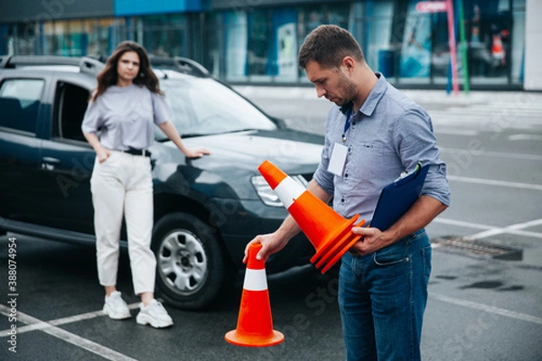 Fototapeta Male tired driving instructor collects traffic cones at the end of the work shift. Blurred female in light casual clothes student at background standing near the black car. Wet asphalt after rain.