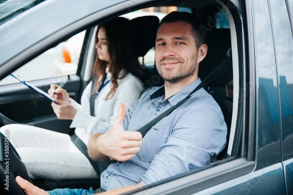 Fototapeta Happy male student showing thumbs up and smiling. Safe driving concept. Cheerful young man excited about his successful driving license exam. Female instructor wrtiting results of test in her paper.