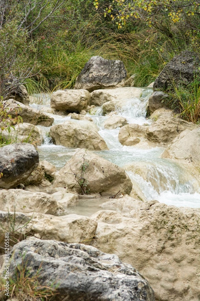 Fototapeta A river in South France, flows between rocks, trees and grasses. Forms little cascades. Early autumnal colors. Picture in portrait mode.