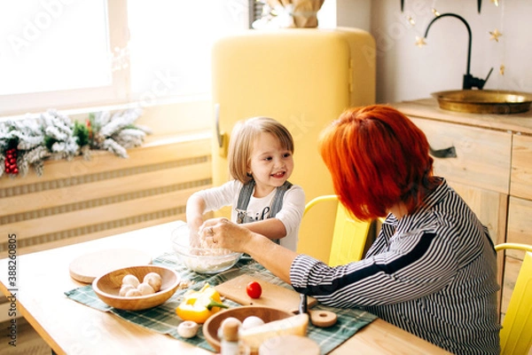 Obraz Cook together at home. Red-haired grandmother and little granddaughter cook pizza in a cozy home. Have fun in the kitchen
