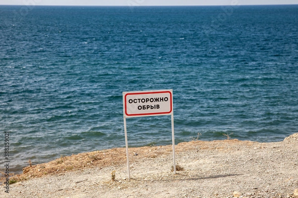 Fototapeta Sign "Caution cliff" at the edge of the cliff in the sea
