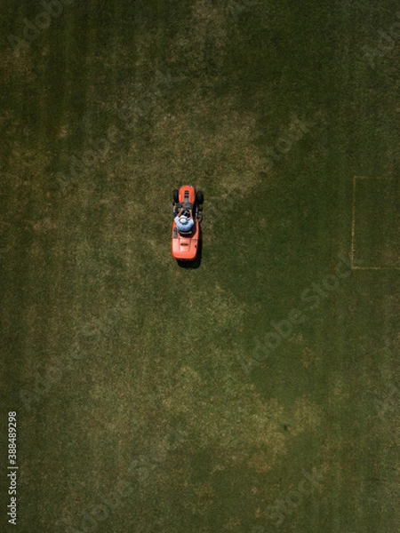 Fototapeta Aerial top down view of man mowing grass on football stadium. Red mower machine and driver. Minimalism. Art drone photography.