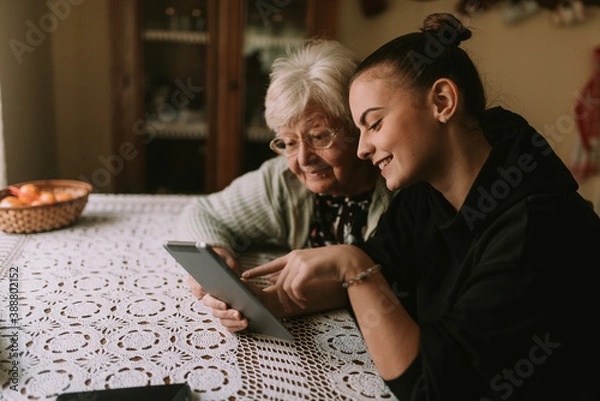 Fototapeta A smiling caucasian grandmother and her teenage granddaughter look at a tablet while sitting at a table in the kitchen. The granddaughter teaches her grandmother to use new technology