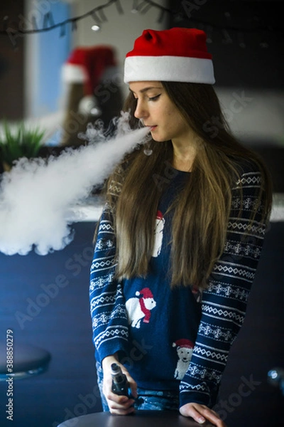 Fototapeta Vape teenager. Young pretty white caucasian girl in a hat of Santa Claus smoking an electronic cigarette in vape bar. Bad habit that is harmful to health. Vaping activity.