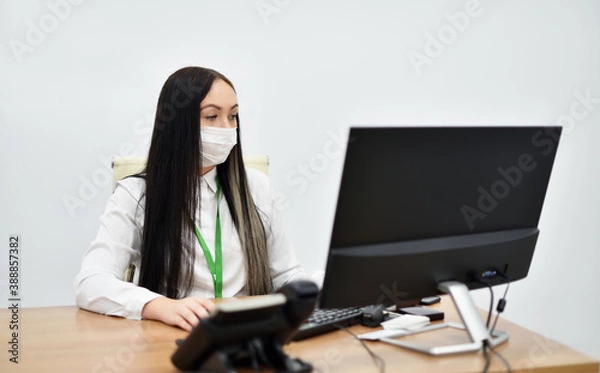 Fototapeta A young female Bank employee in a face mask works with a desktop computer in the office