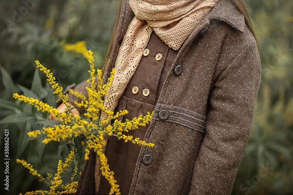 Fototapeta A little girl in a coat and a brown linen dress on a background of autumn leaves. The yellow autumn  flowers in the hands of the baby girl. Autumn time. Image with selective focus
