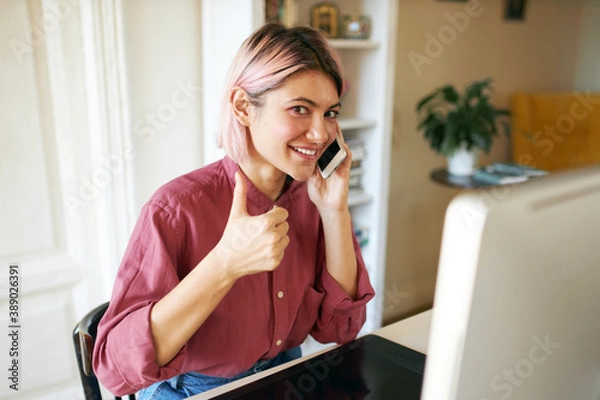 Fototapeta Cute young woman designer with pink bob hairstyle working from home, sitting at table with computer and graphic tablet, having phone conversation, showing thumbs up gesture at camera and smiling