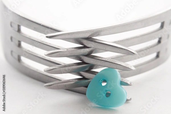Obraz Closeup of the crossed tines of two forks. Next to it is a blue button in the shape of a heart . White background, selective focus
