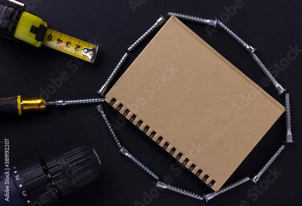 Fototapeta tools for repair and construction. Screwdriver, measuring tape, furniture screws, notebook on a black background with a place for text. View from above